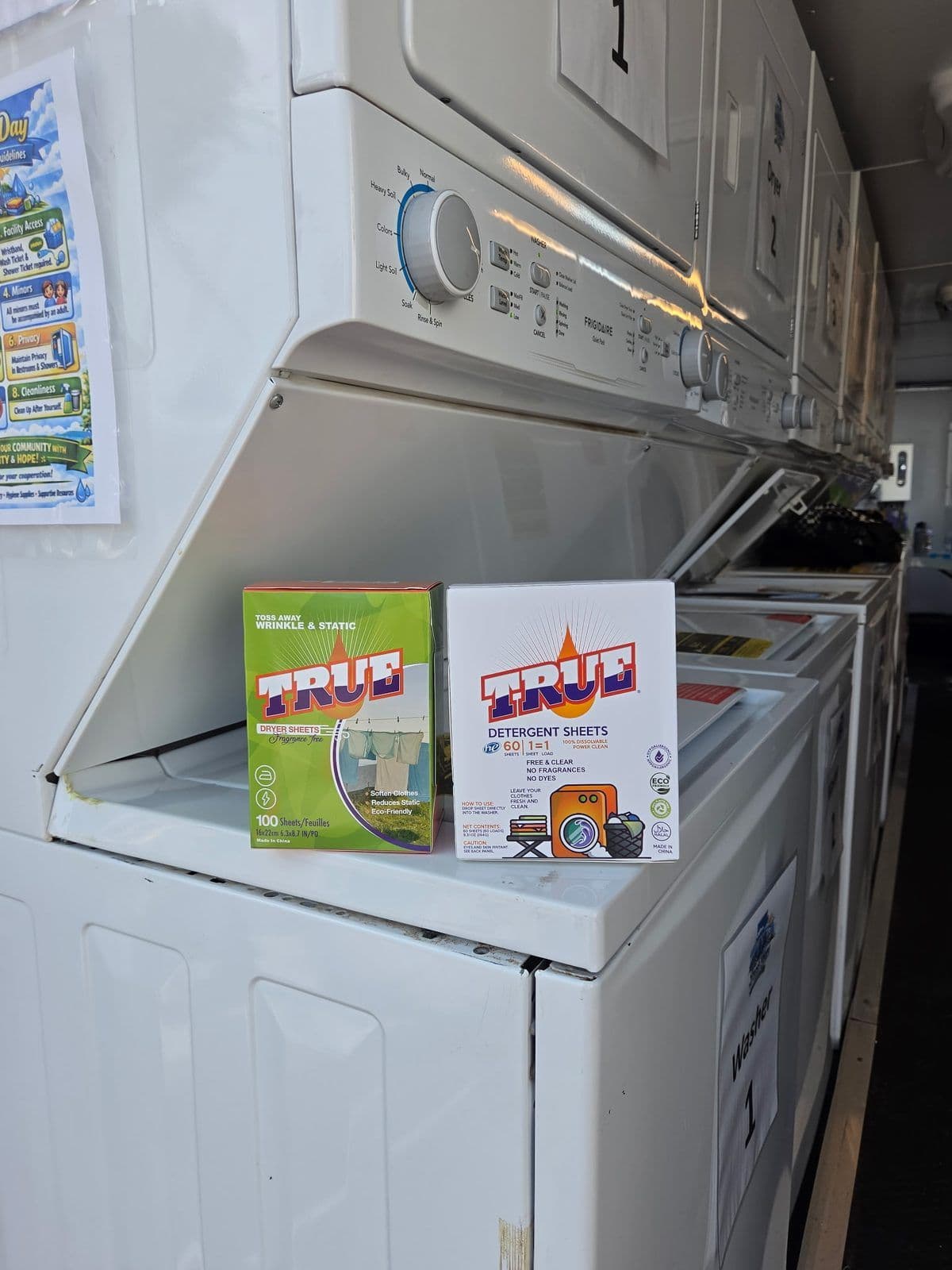 Washers and dryers lining the inside of the Fresh Hope laundry trailer, with boxes of True detergent sheets and dryer sheets set out on an open machine lid.